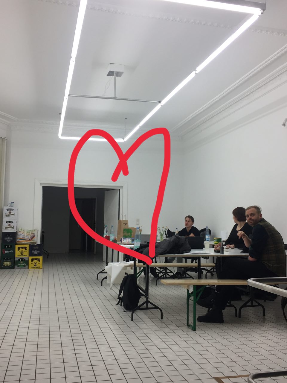 A bright office space with strip lighting and people seated at tables, overlaid with a large red hand-drawn heart annotation