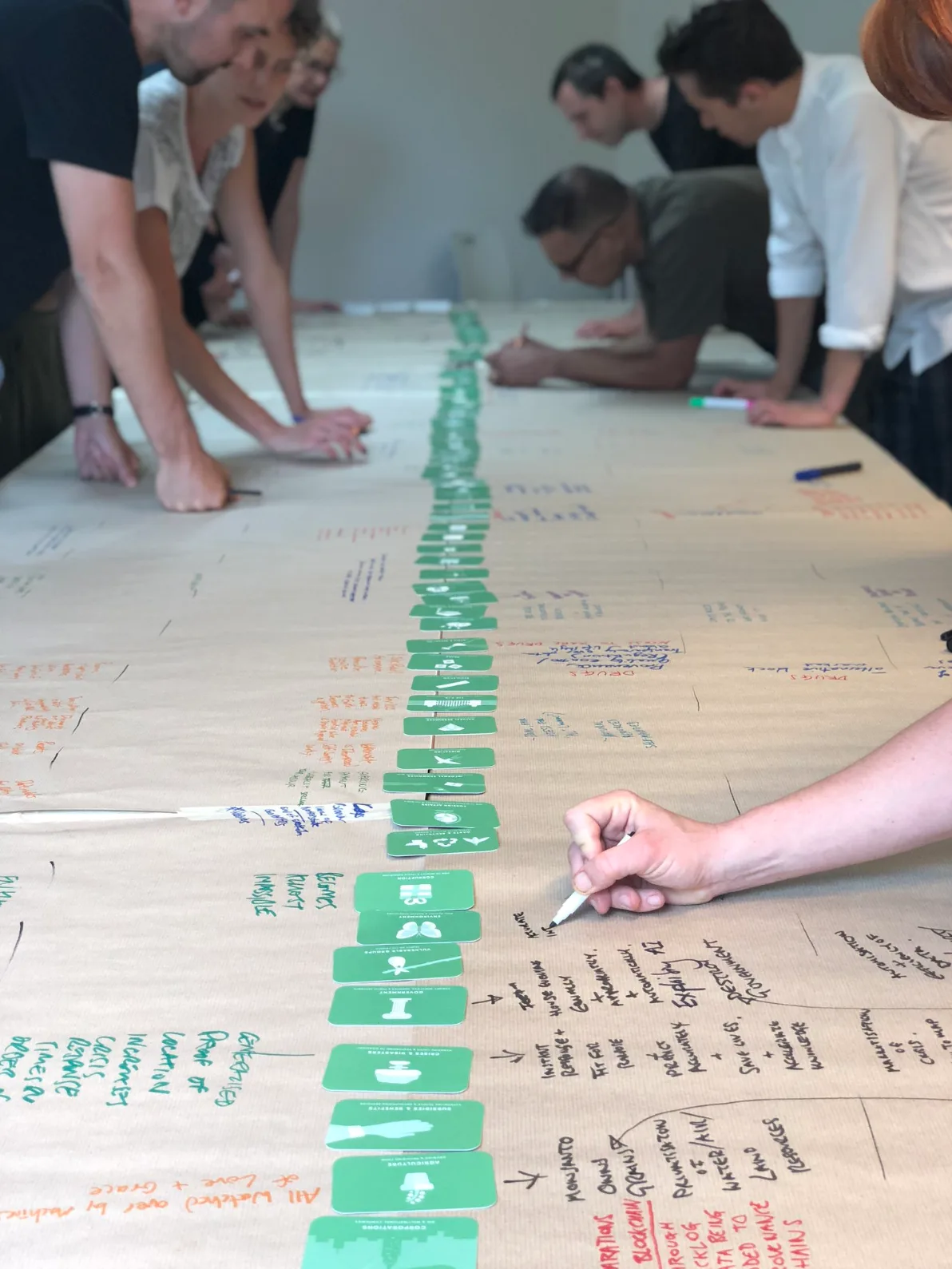 Group of people leaning over a large paper diagram covered with green sticky labels and handwritten notes during a collaborative workshop