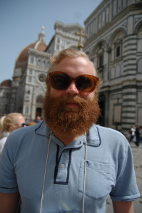 Bearded man in sunglasses taking a selfie in front of Florence Cathedral with its striped marble facade and dome visible behind him