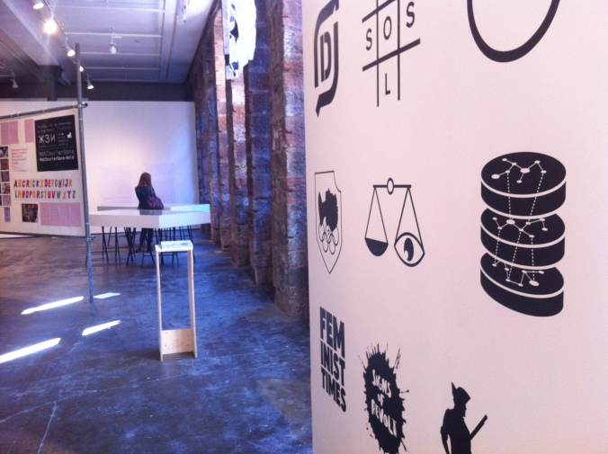 Exhibition space with a large pink wall featuring bold graphic icons including scales, stacked coins, and Feminist Times text; a person seated at a table in the foreground beside exposed brick columns