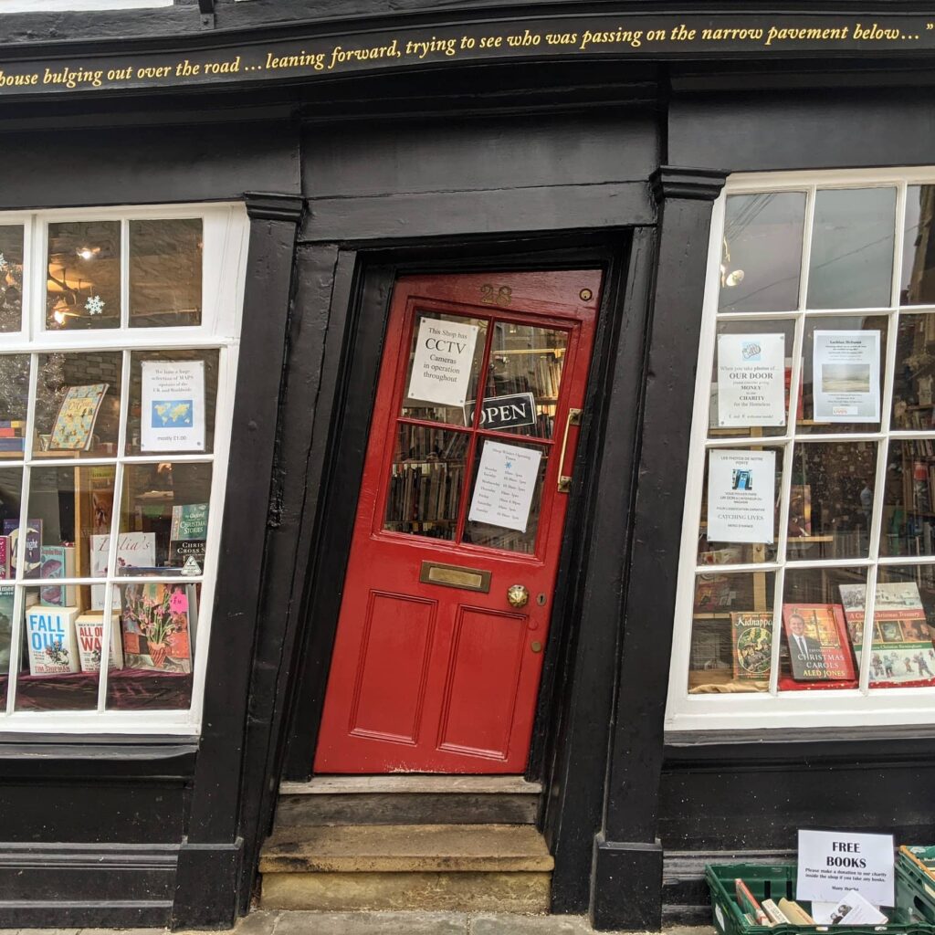 Bookshop storefront with crooked bright red door marked "OPEN", display windows with books and posters on black facade