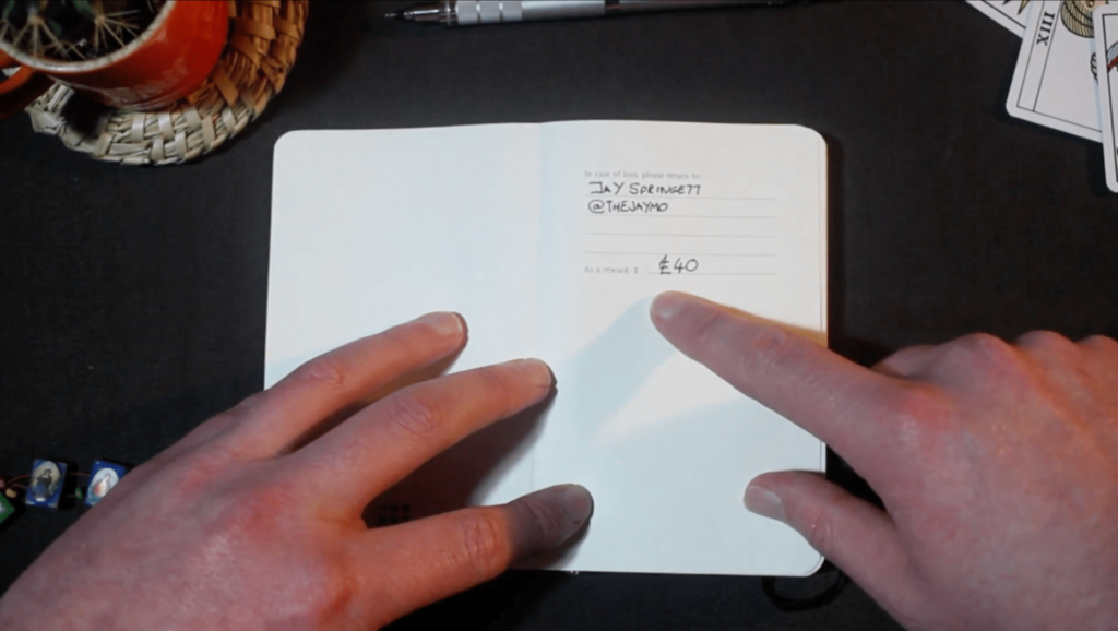 Two hands pointing at a handwritten notebook page with text and a reward amount