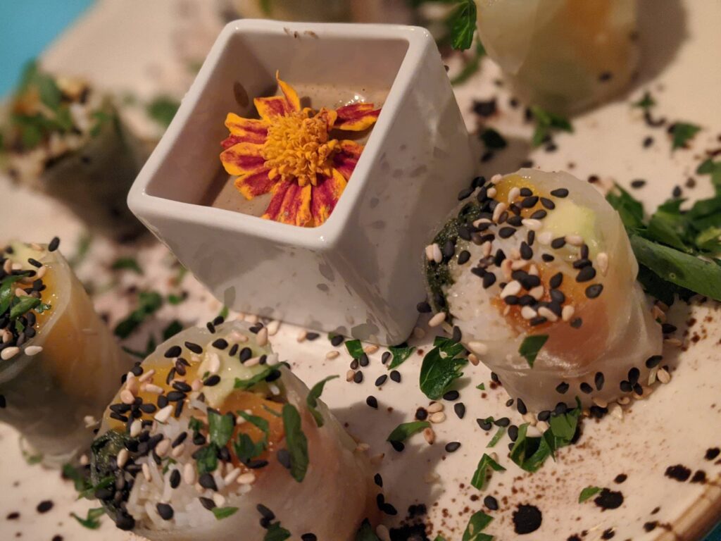 Sesame-topped food items in sand with white ceramic dish containing an orange and pink flower