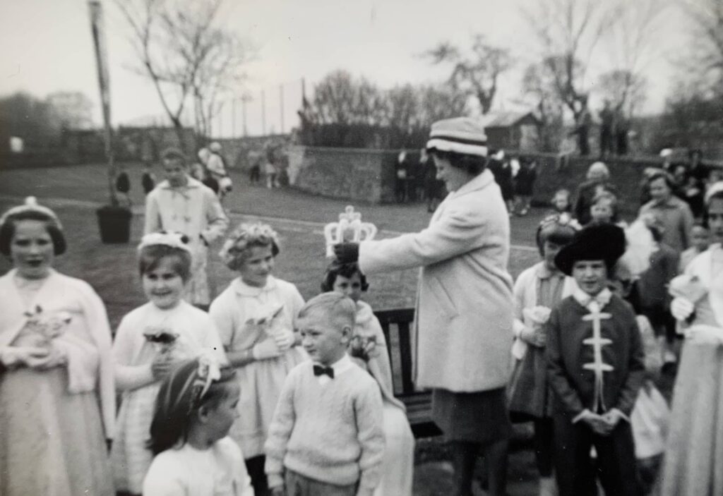 Black and white vintage photograph of children gathered outdoors with an adult man in a hat conducting or addressing the group in an open field with bare trees in the background