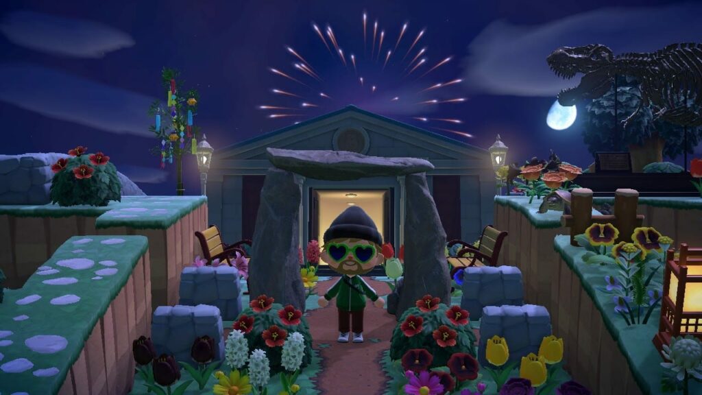 Animal Crossing screenshot of nighttime holiday scene with character in green standing before a building surrounded by colorful flowers and fireworks under full moon