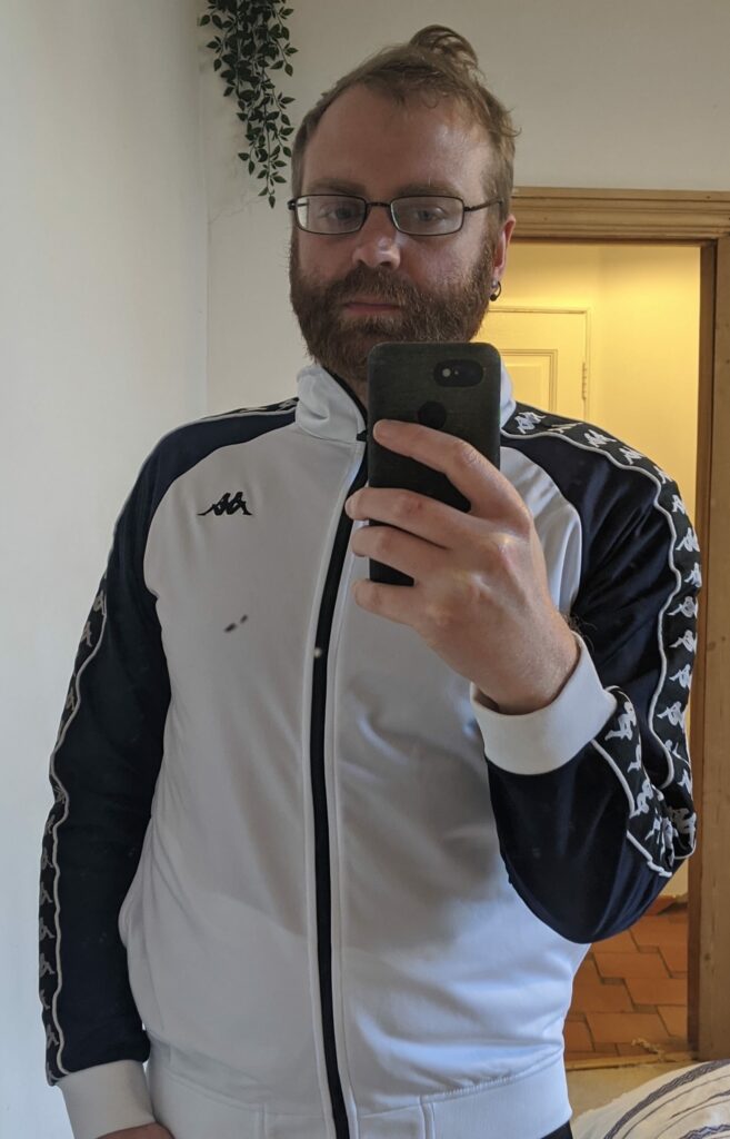 Selfie of person wearing glasses and black and white Kappa tracksuit, holding phone for mirror selfie indoors