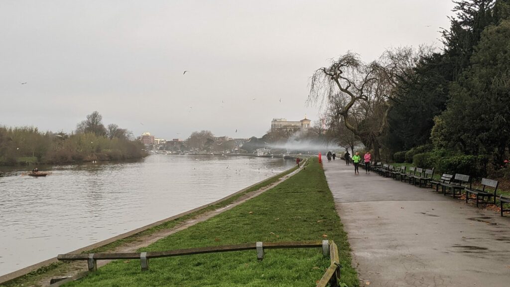 Riverbank path with wooden benches beside a wide river with bare trees and people walking in winter light
