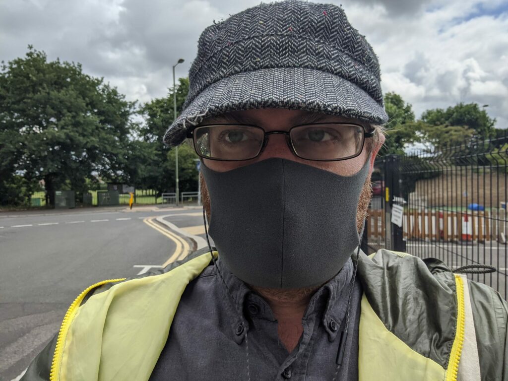 Man wearing glasses, face mask, patterned hat and yellow safety vest outdoors