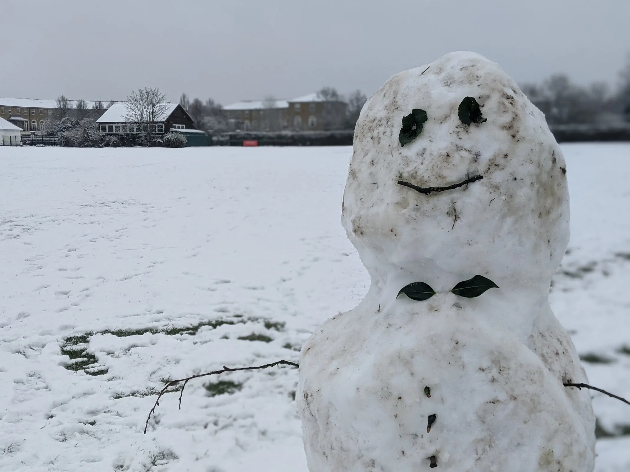 Smiling snowman with coal eyes and mouth on snowy frozen lake with houses and trees in background