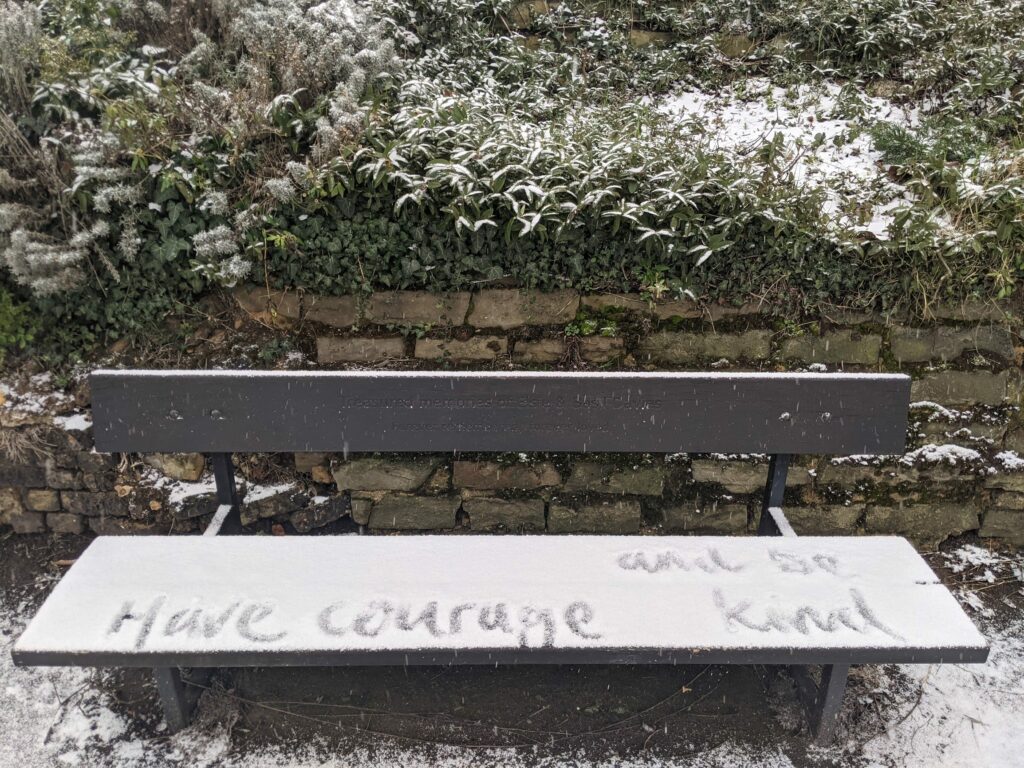 Wooden bench with handwritten text "Have courage" and "Live" surrounded by green foliage and ivy