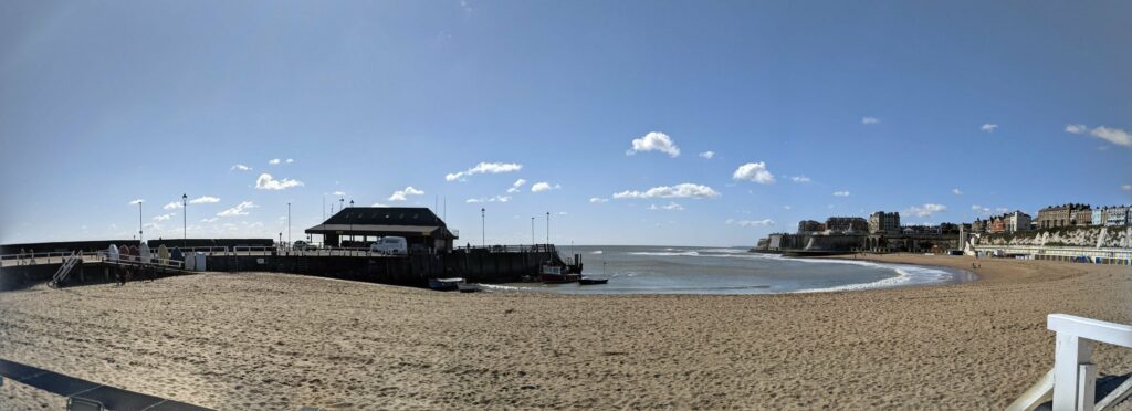 Panoramic view of a beach with sandy shore, pier structures, buildings along the coastline, and blue sky with clouds