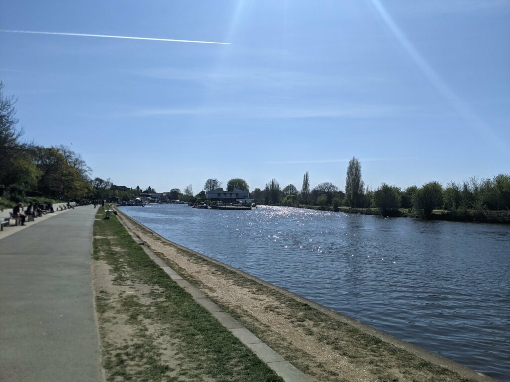 Riverside path with paved walkway, green grass, blue water, trees and buildings along the bank under clear sky