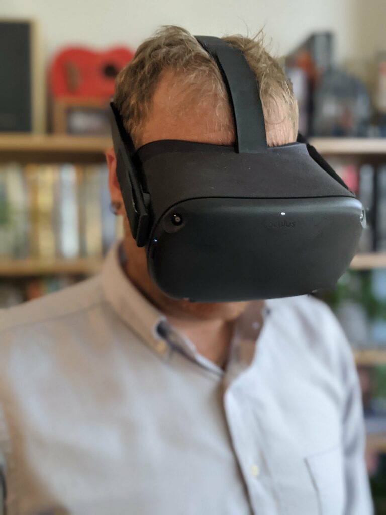 Person wearing black VR headset in white shirt indoors with blurred bookshelf background