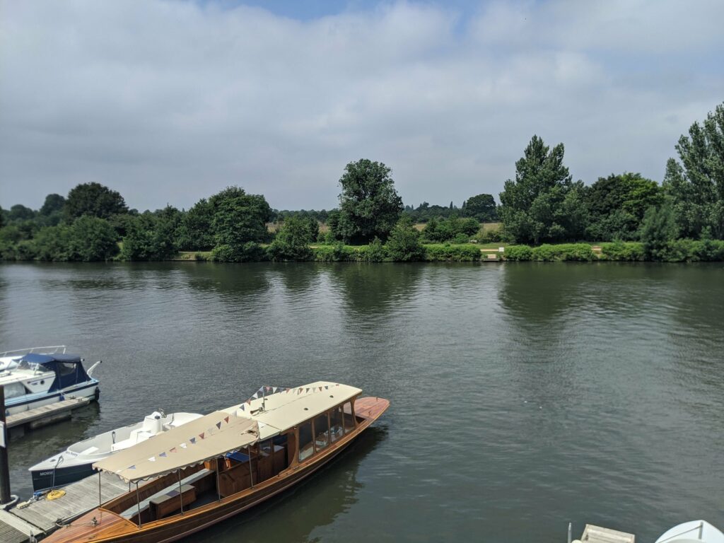 Wooden riverboat with blue cover moored on calm river, additional smaller boat visible to the left, tree-lined banks in background