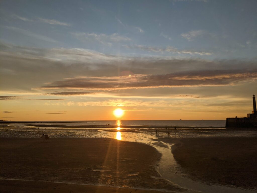 Sunset over beach with golden sun reflecting on wet sand and shallow water, dramatic orange and blue sky