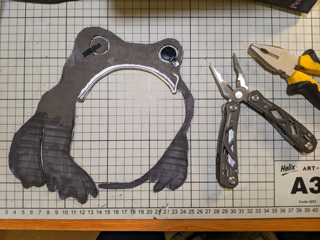 Dark puppet head with two circular eyes and white-outlined mouth beside pliers and yellow-handled tool with ruler on tiled background