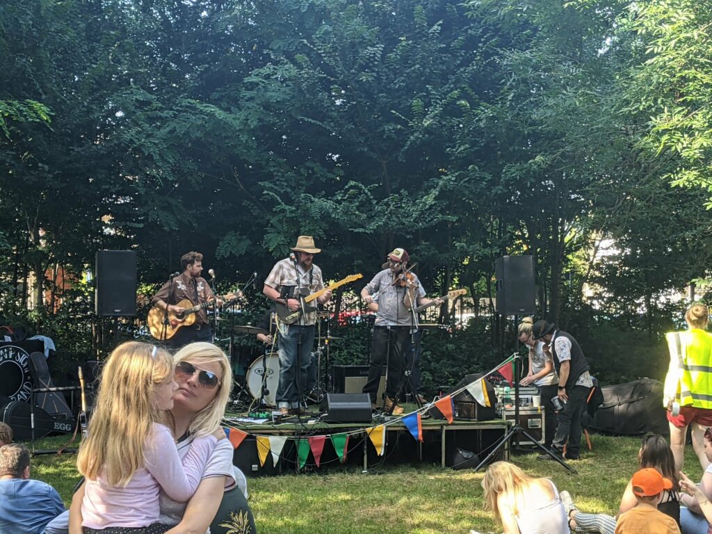 Outdoor music festival with band performing on small stage among trees, audience sitting on grass wearing sunglasses