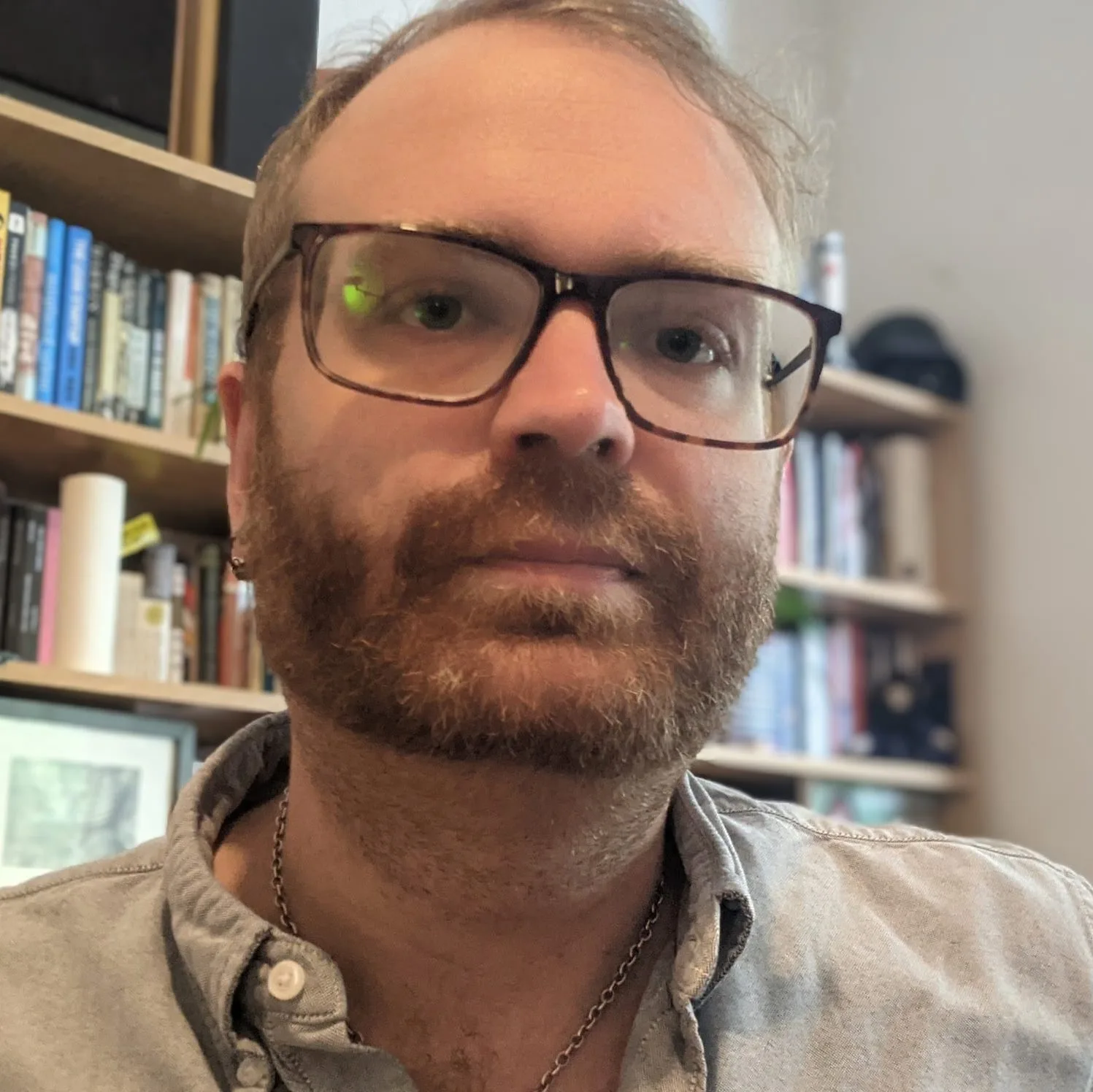 Portrait of bearded man with glasses wearing tan jacket and chain necklace indoors with bookshelves in background
