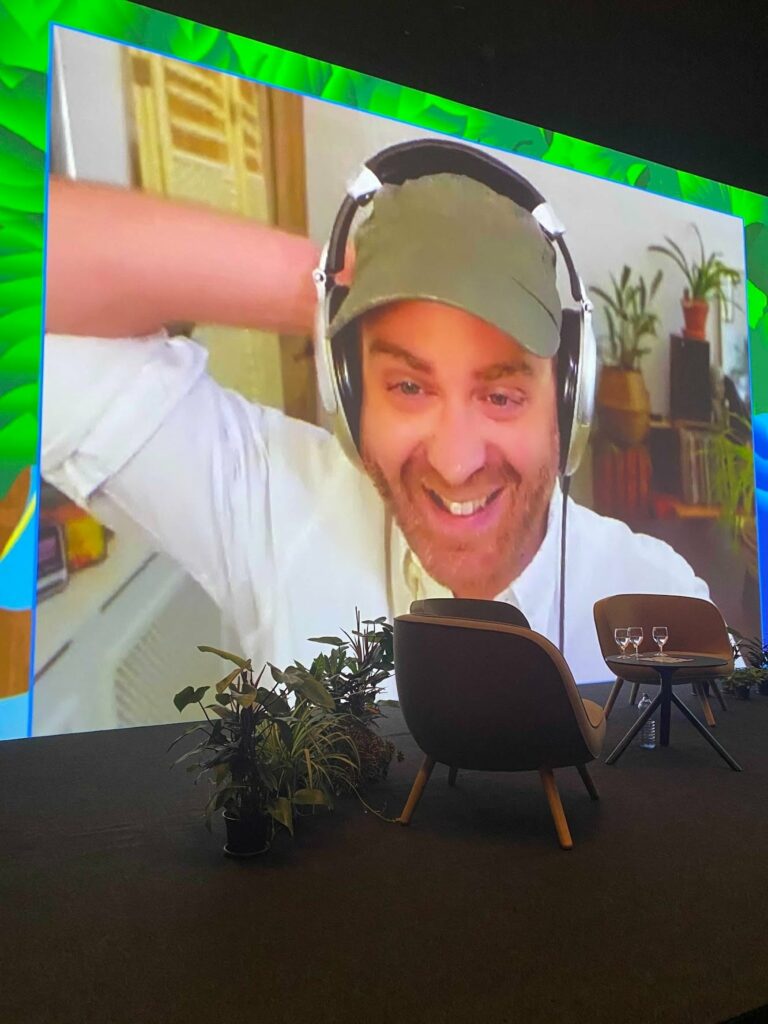 Person wearing headphones at broadcast studio desk with large monitor showing video call participant in background