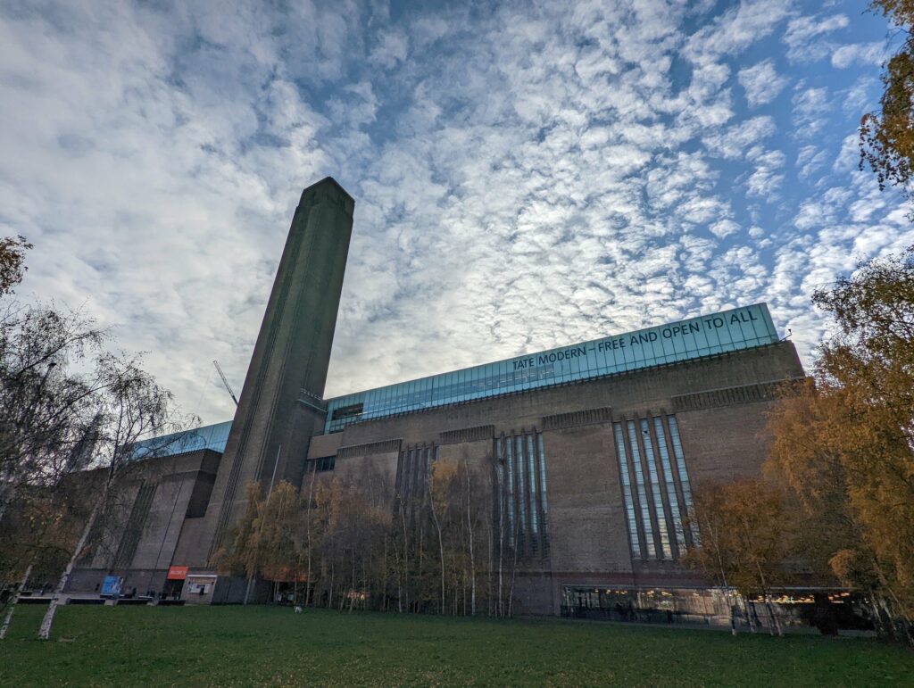 Tate Modern museum with distinctive brick tower and modern blue glass extension, green lawn and autumn foliage
