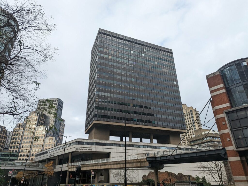 Brutalist high-rise office building with grid-pattern facade and concrete structure, connected by overhead bridge, against overcast urban skyline with bare trees