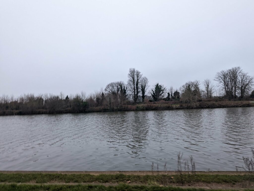 River or pond with still water and bare winter trees reflected on the bank under overcast grey sky