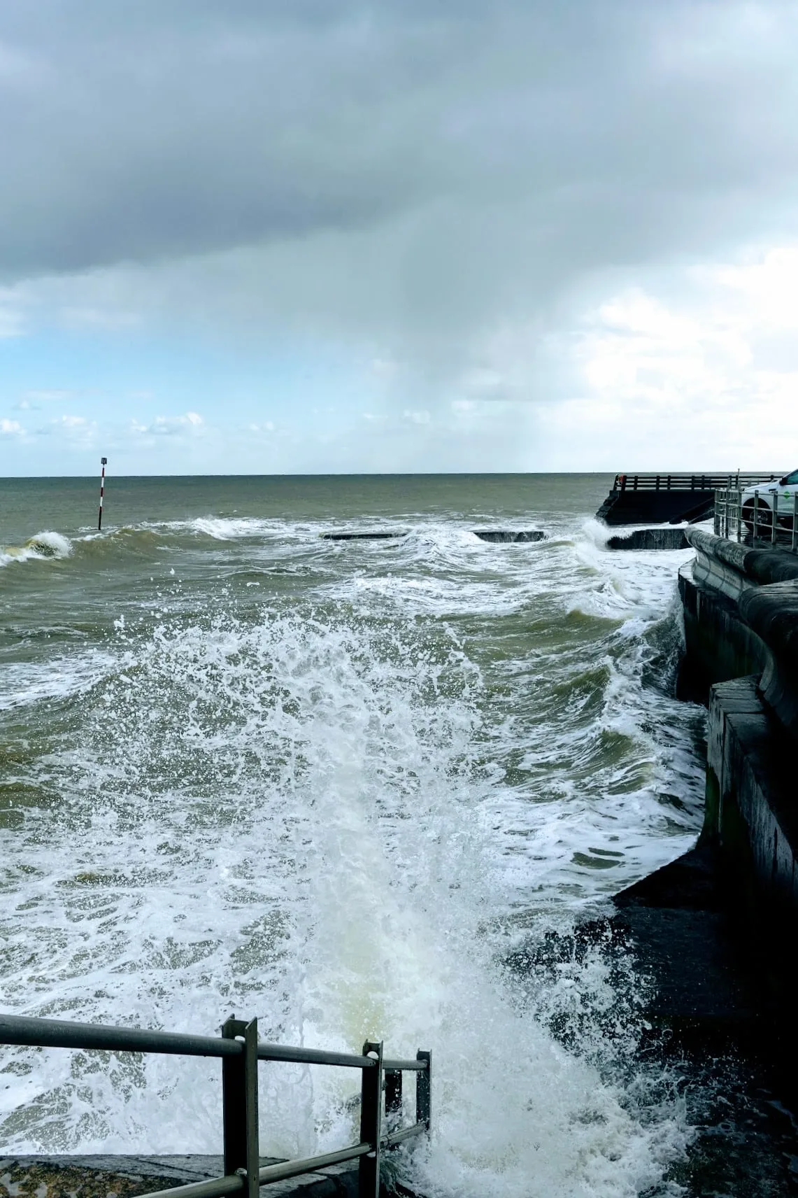 Rocky sea wall with foaming white waves breaking on rocks, stormy greenish sea, navigation buoy marker in distance