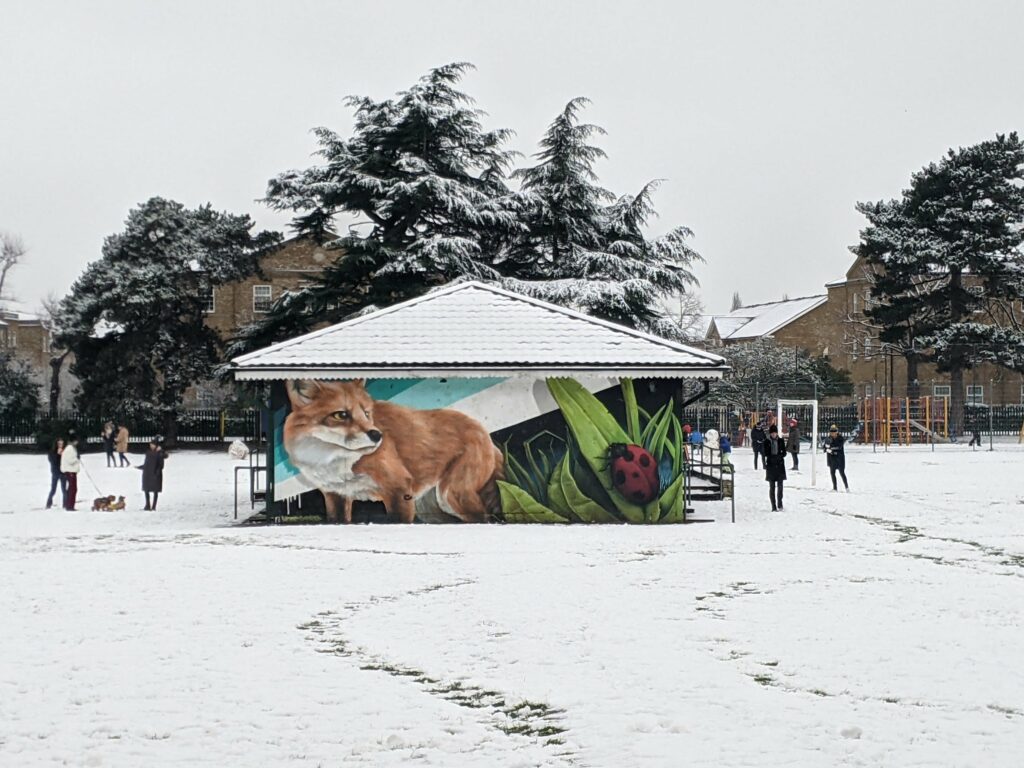 Snowy beach with a building pavilion decorated with artwork of a fox and plants, people walking on snow-covered sand, trees in background