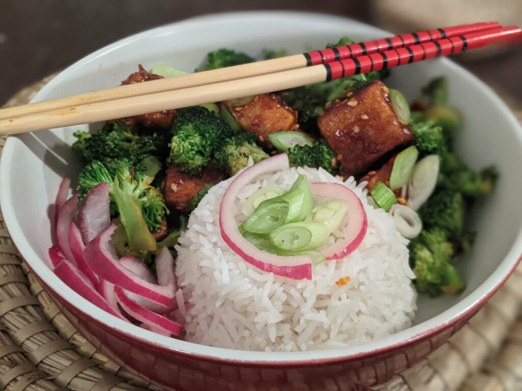 Bowl of Asian cuisine with white rice, broccoli, glazed meat cubes, cucumber slices and radish, held with red and wooden chopsticks
