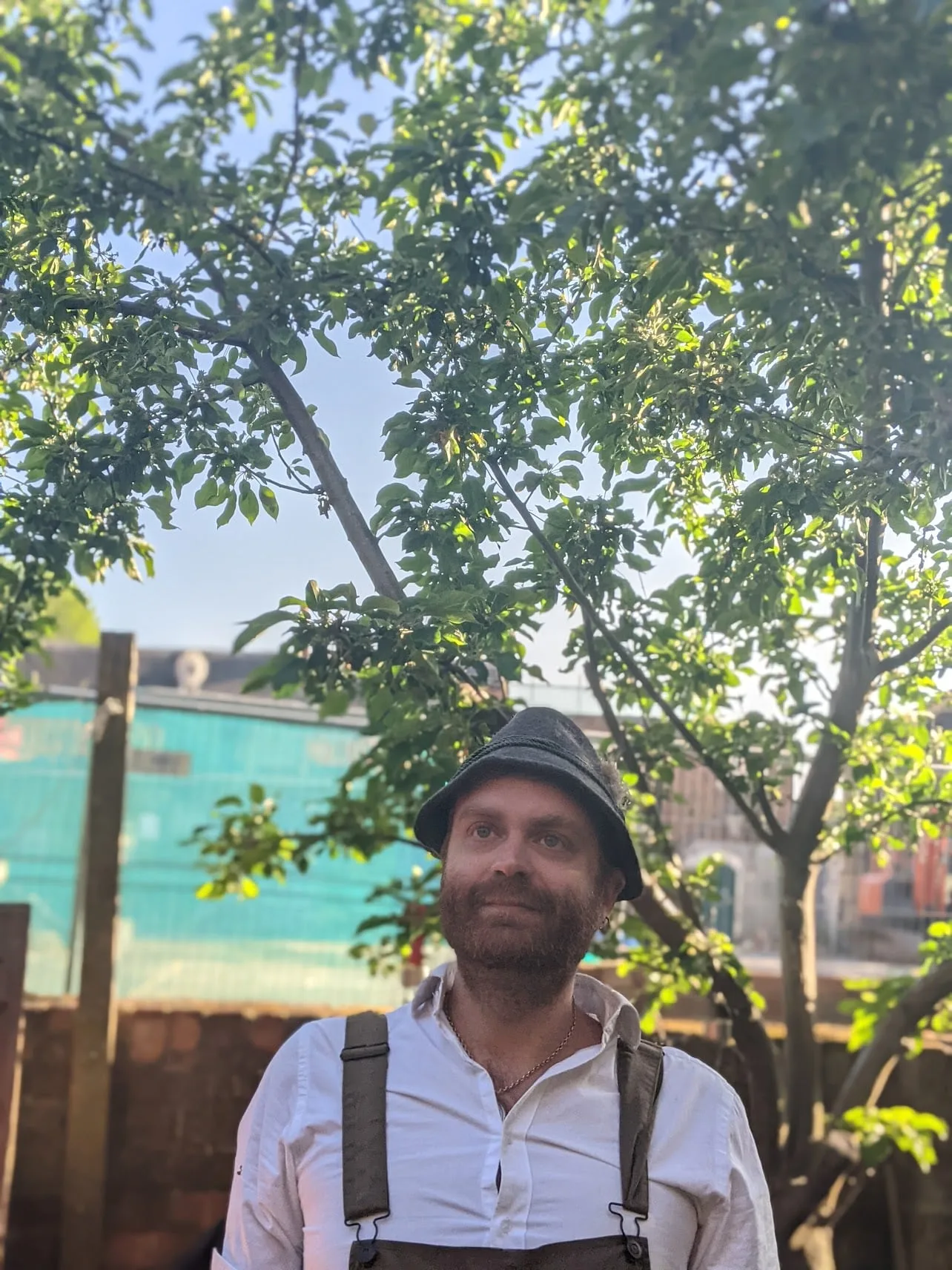 Person with beard wearing black cap and white shirt with backpack straps, standing beneath leafy tree with blue sky, outdoor structure visible