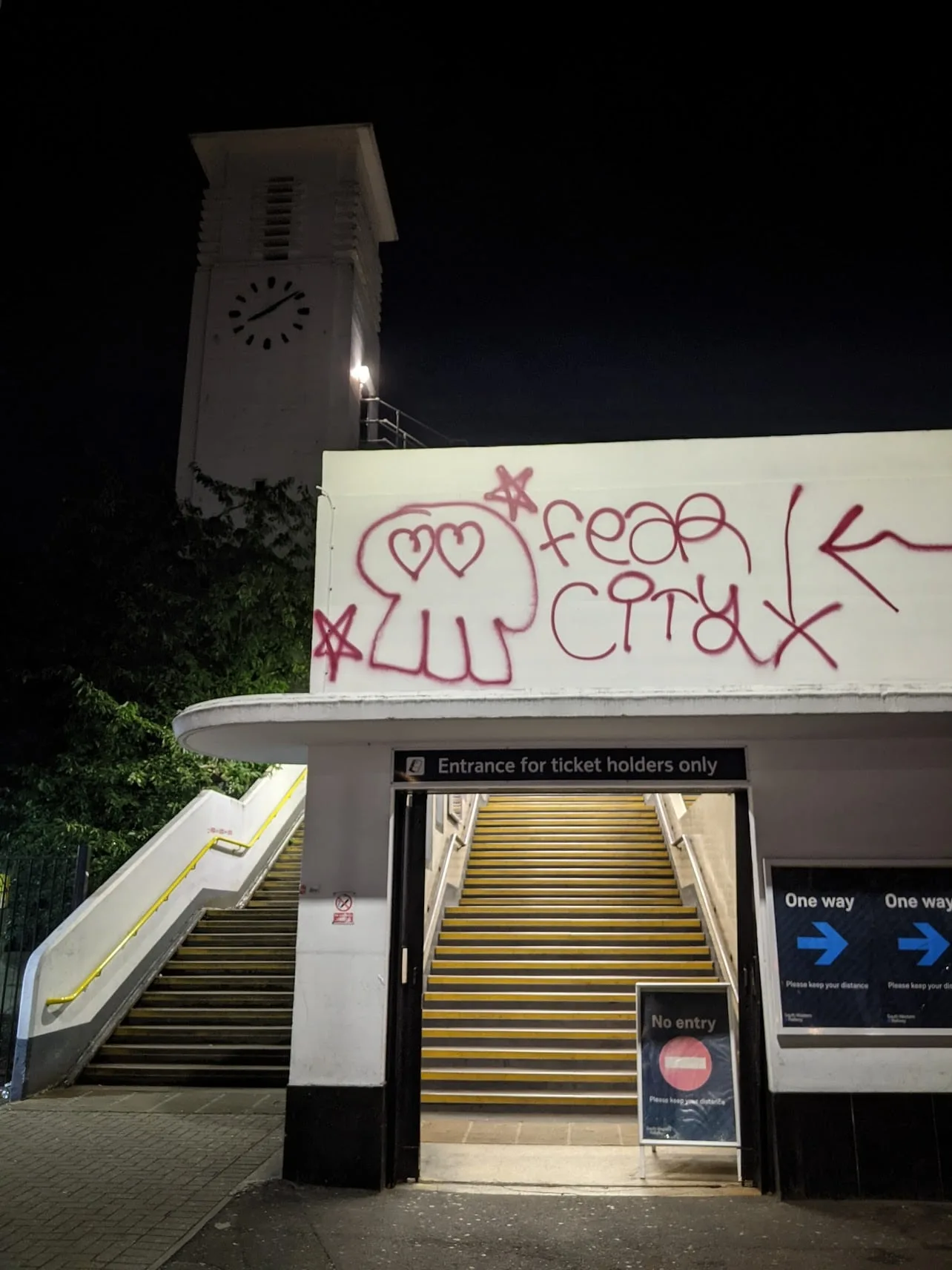 Night shot of waterfront building with red graffiti on white sign, stairs with yellow handrails, clock tower above, "Entrance for ticket holders only" sign