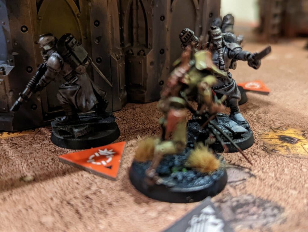 Warhammer 40K tabletop game scene with miniature soldier figures on terrain with red orange game markers