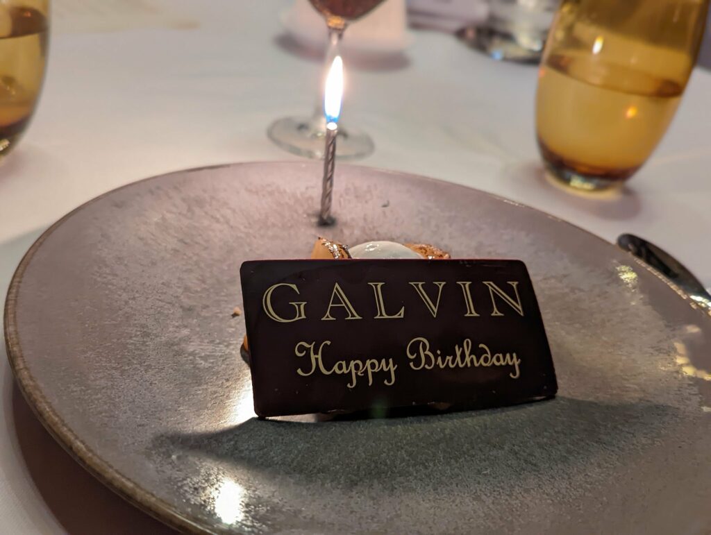 Birthday dessert with dark chocolate plaque reading "GALVIN Happy Birthday" on grey plate with beer bottle and candle in background