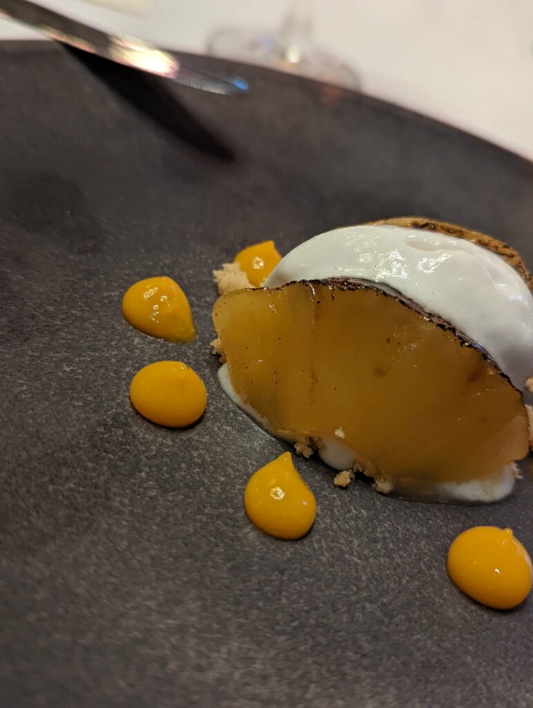 Fine dining dessert plated on dark slate with a baked main component topped with white foam and golden spheres arranged on the plate