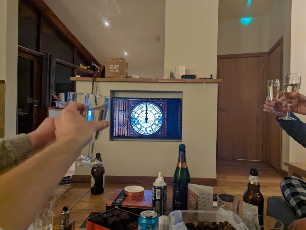 People toasting with glasses at a New Year's Eve celebration, with bottles and food on a wooden table, a blue and white clock display on the wall
