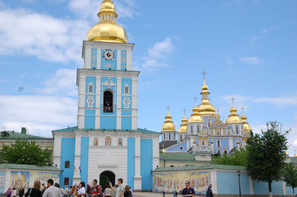 Historic religious complex with blue and white bell tower, golden domes, and crosses under blue sky with visitors gathering