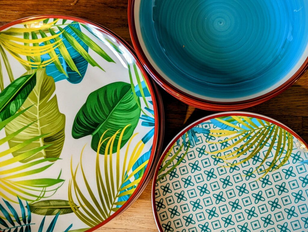 Overhead view of colorful decorative dishes on wooden surface: white plate with tropical banana and foliage design, turquoise bowl, and patterned bowl with geometric crosses and squares
