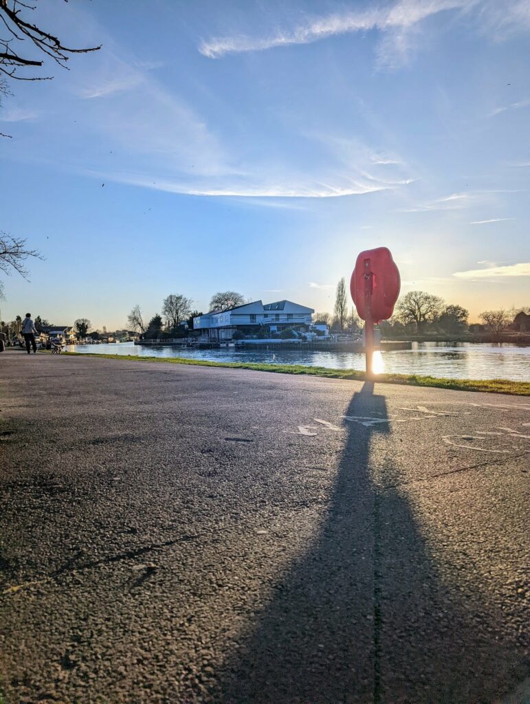 Red balloon sculpture above pavement at riverside park during golden hour, with blue building and people visible in background