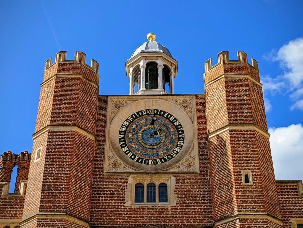 Red brick historic building with twin crenellated towers and central tower with large clock face, blue sky background