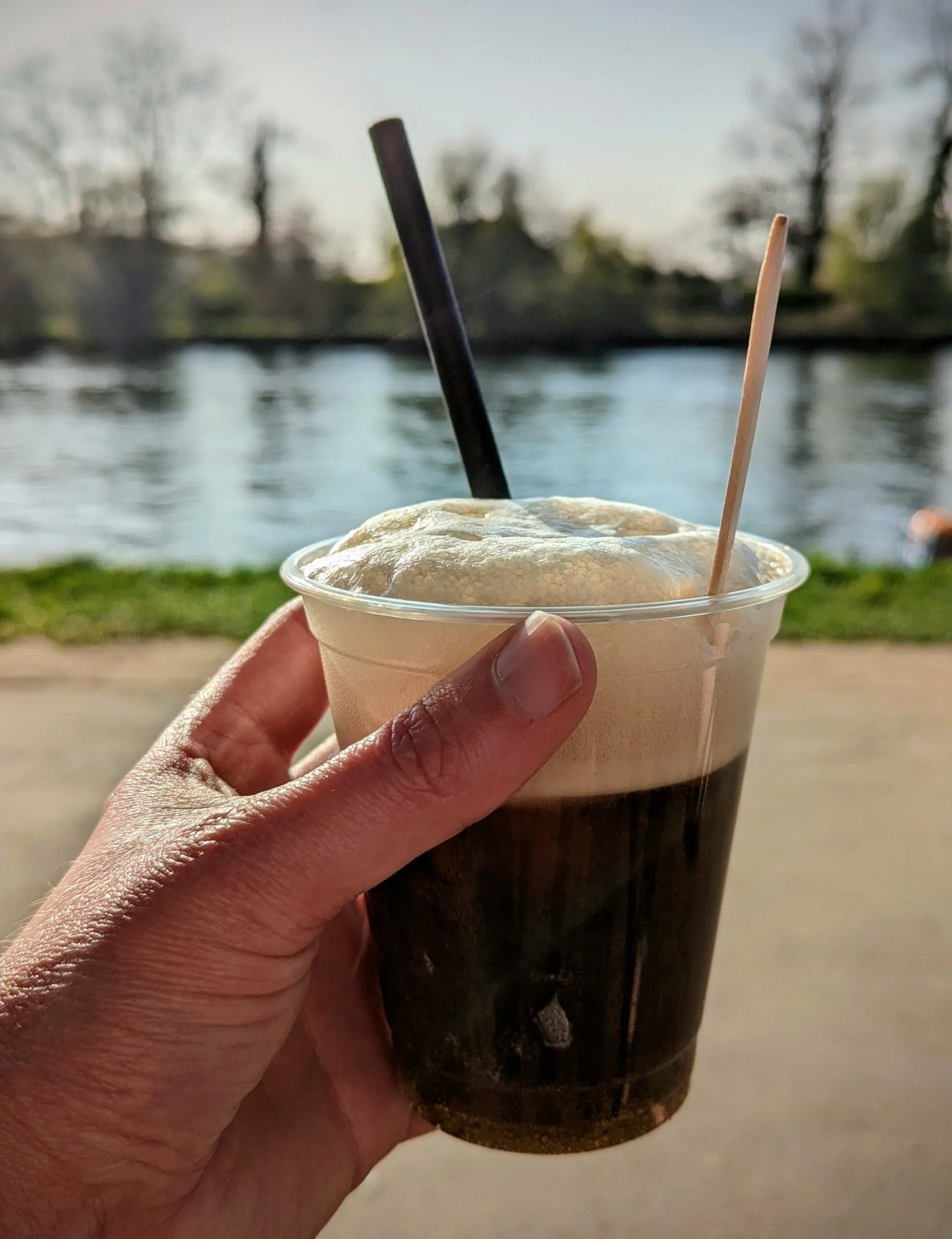 Hand holding iced drink by river with trees in background