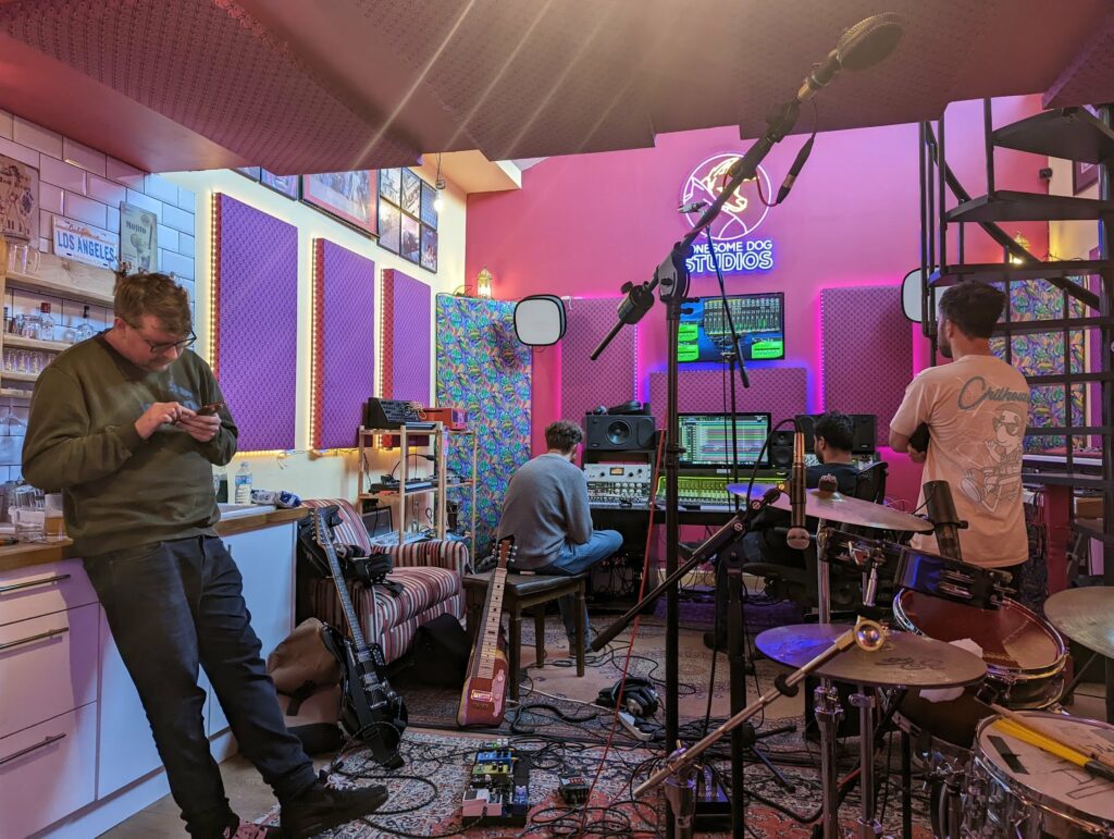 Interior view of Lonesome Dog Studios with neon pink lighting, multiple musicians and producers working, keyboards, mixing equipment and shelving visible