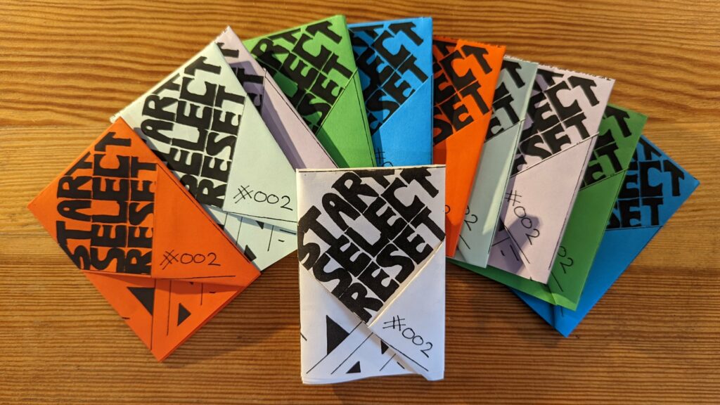A fan of colorful folded zines on a wooden table, each printed with bold black START SELECT RESET text and the handwritten number #002.