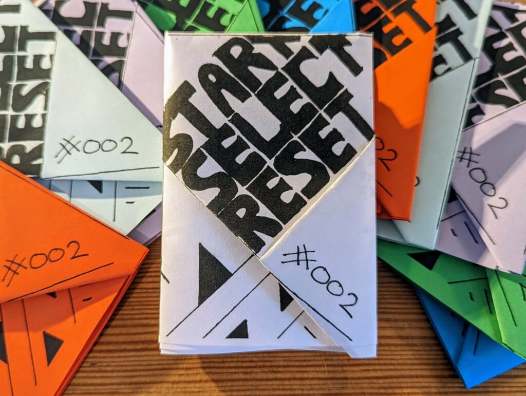 A white folded zine titled START SELECT RESET #002, surrounded by similar orange, green, and blue booklets on a wooden surface.