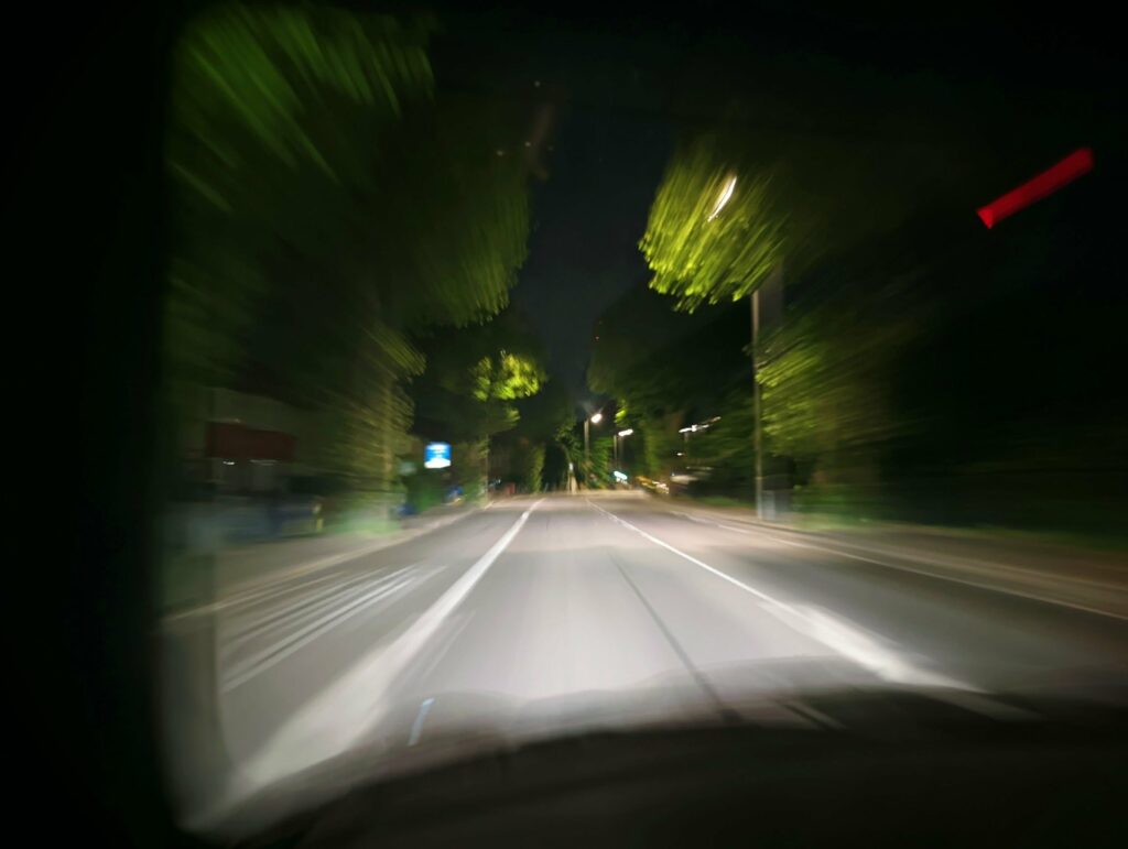Motion-blurred night view from a car traveling down a dark road lined with trees illuminated by headlights, with streaks of white light.