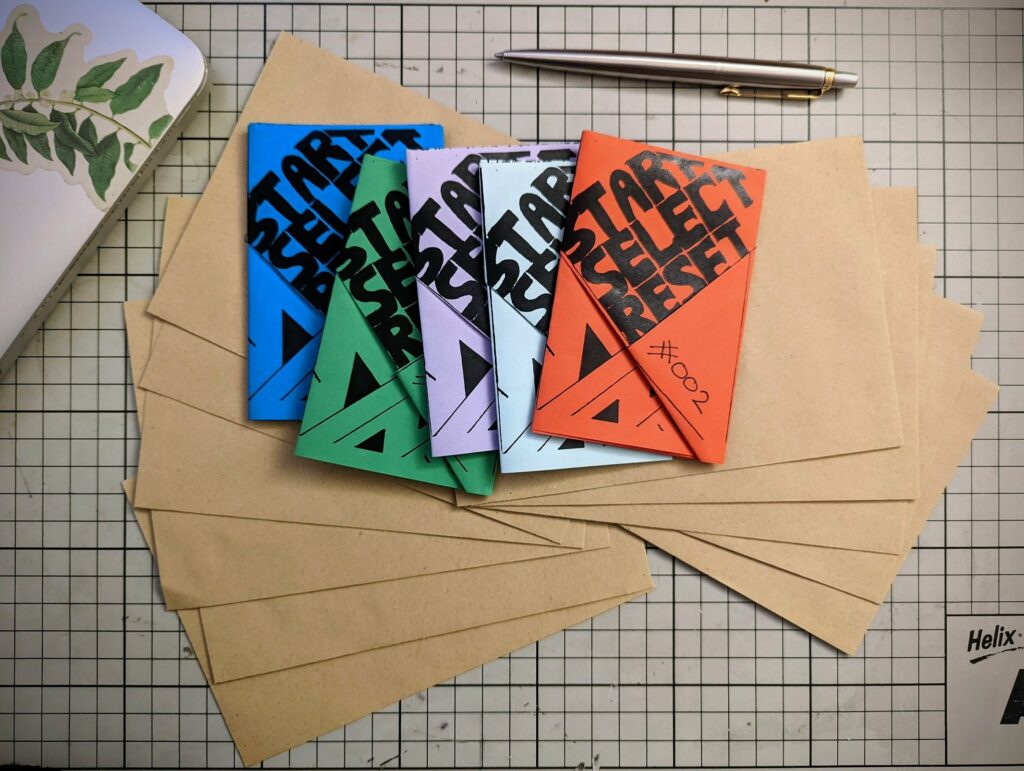 Colorful zines titled START SELECT RESET fanned across brown envelopes on a gridded cutting mat with a silver pen.