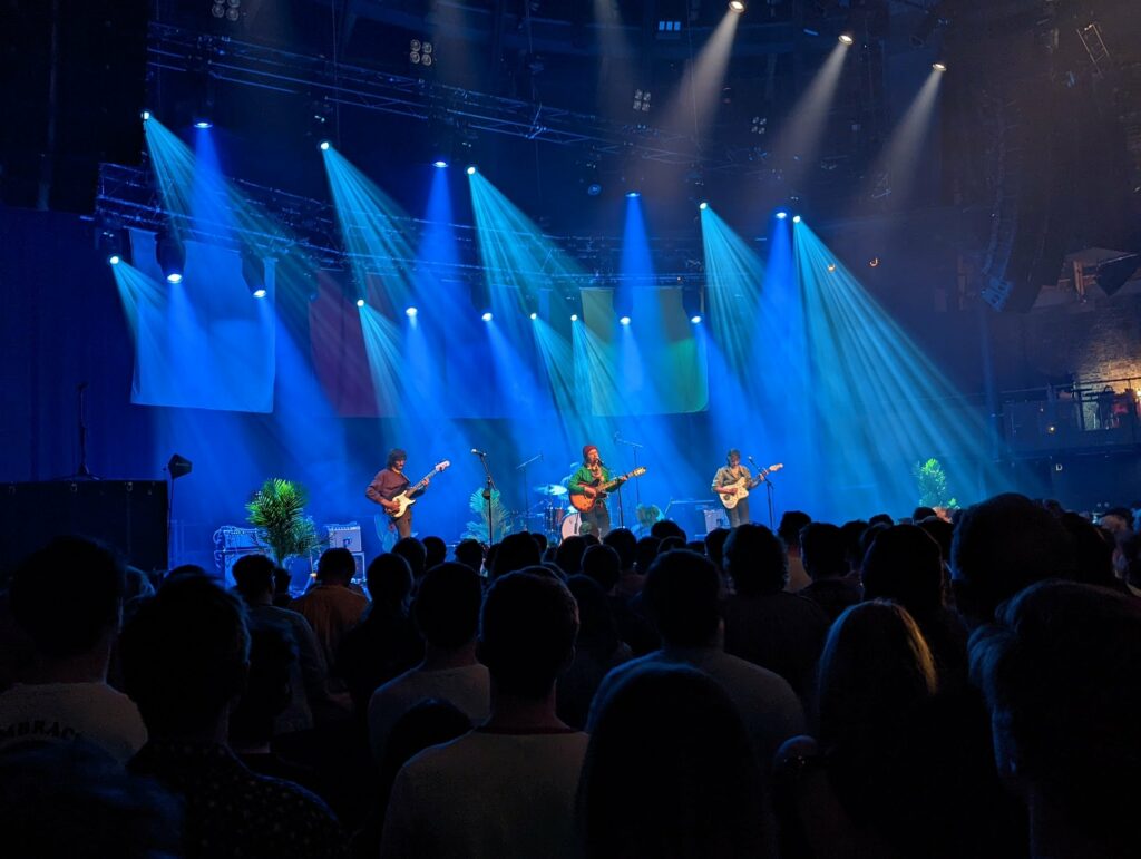 Pinegrove performs on stage under dramatic blue spotlights in a dark concert venue, viewed from behind a silhouetted crowd.