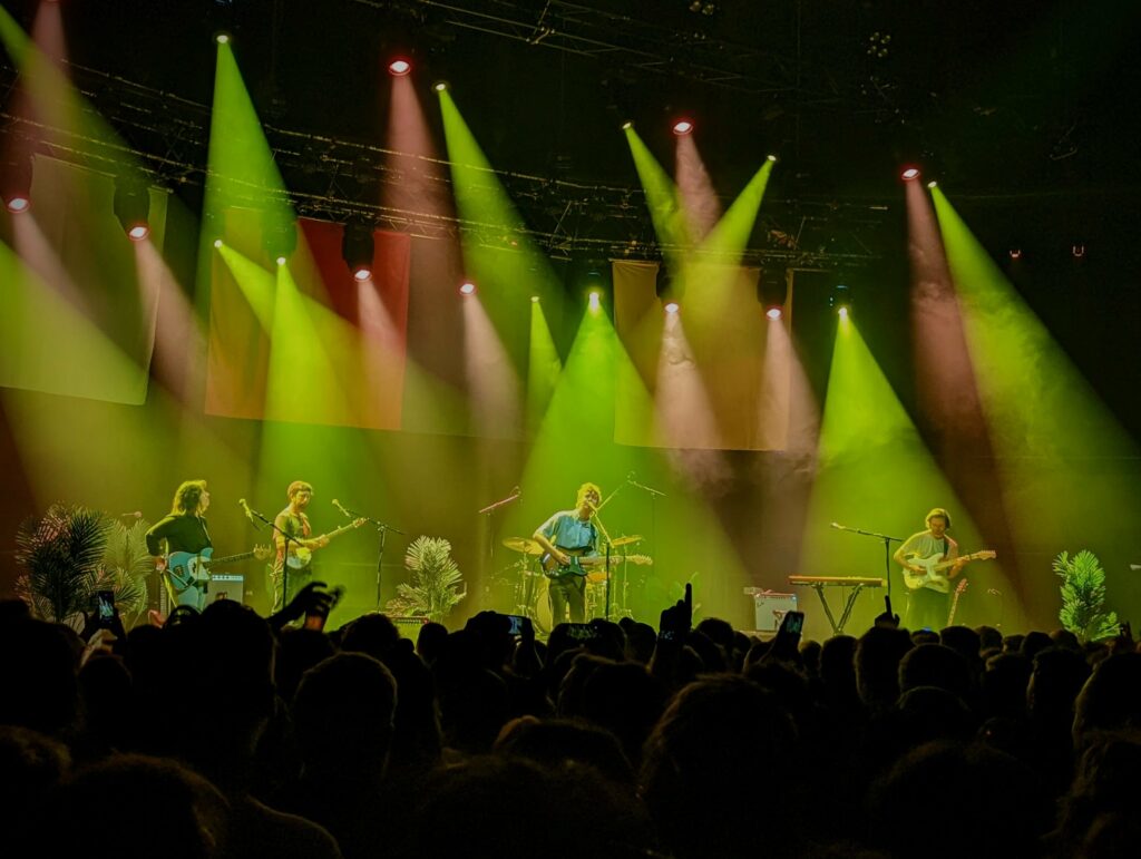 A band performing on stage under vibrant green and pink spotlights, viewed over the silhouettes of a crowd at a live concert.