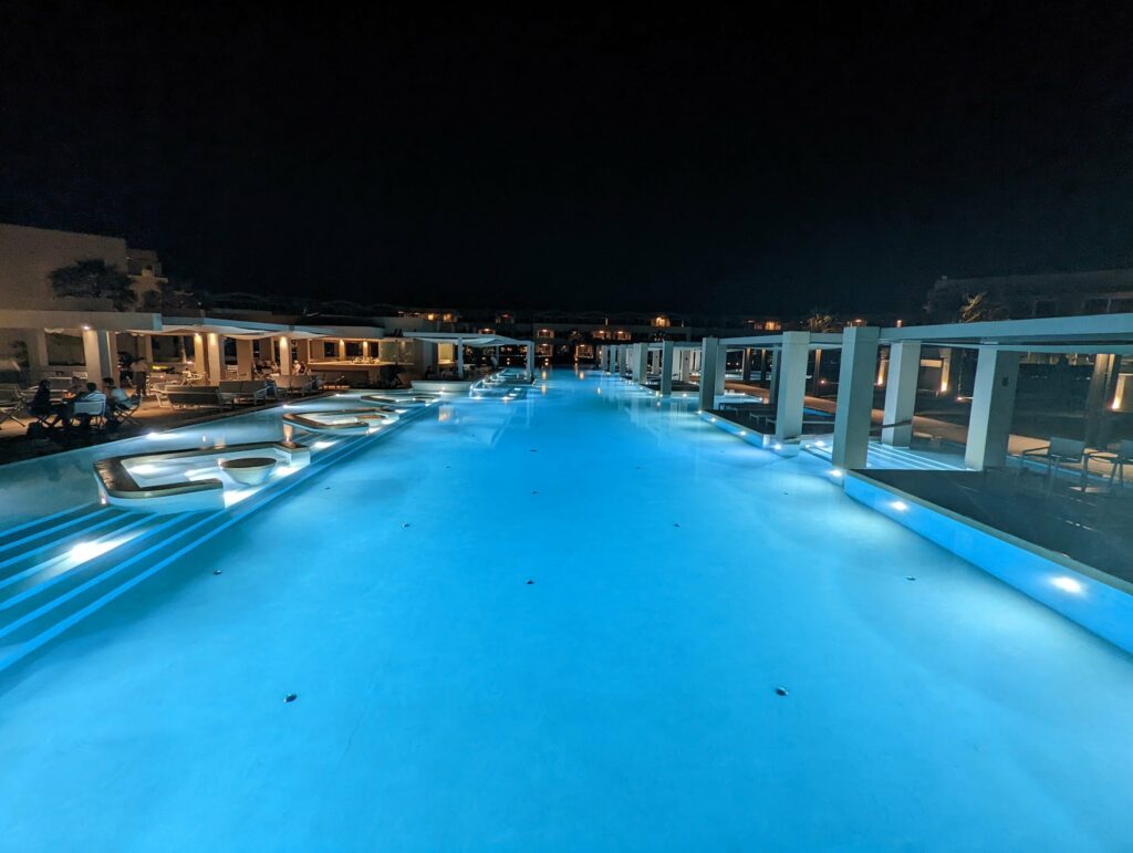 A long, glowing blue swimming pool at night, flanked by modern white resort architecture and poolside seating areas.