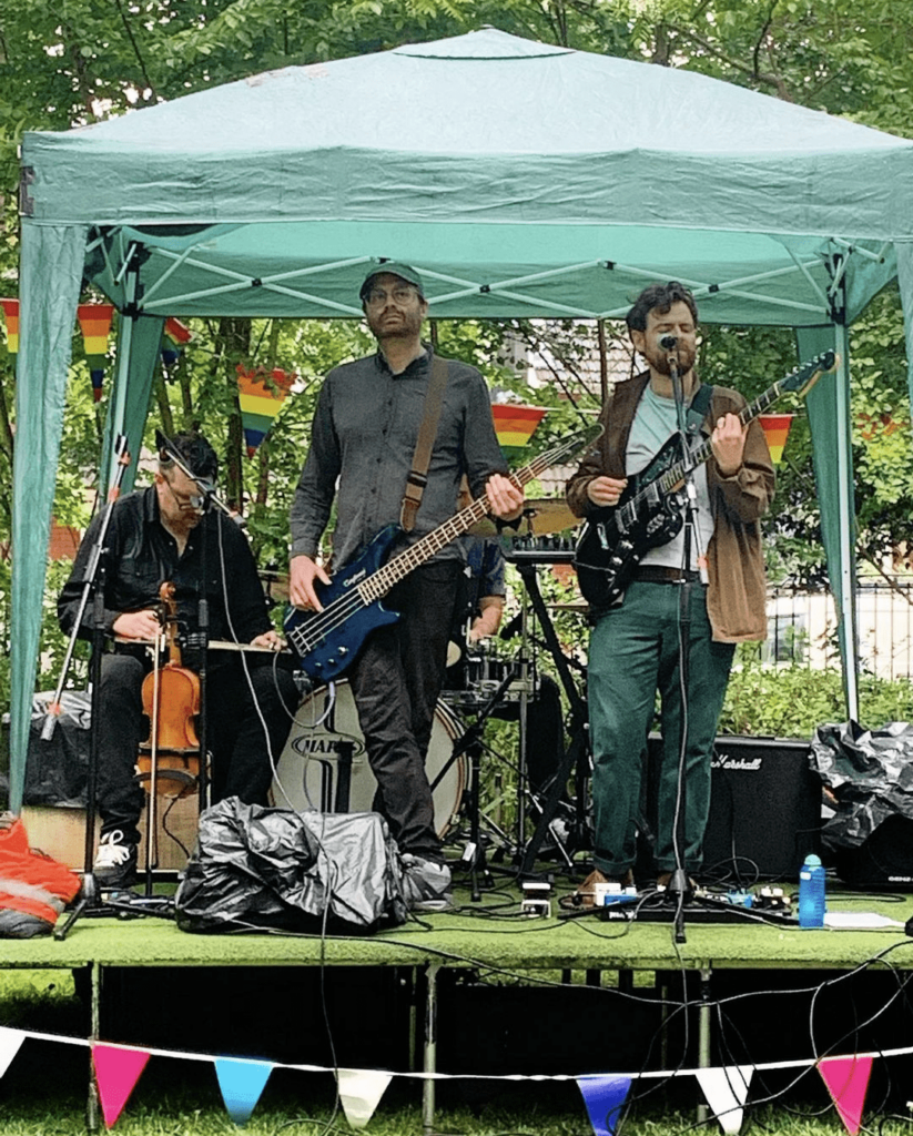The band Forest Bed with a bassist, guitarist, and violinist performs under a green marquee in the rain on an outdoor stage decorated with rainbow bunting.