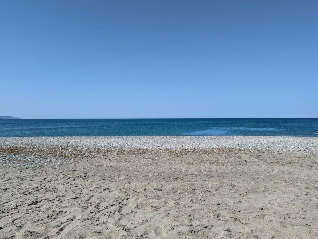 A wide sandy beach meeting a band of grey pebbles before the dark blue sea and a clear, pale blue sky.